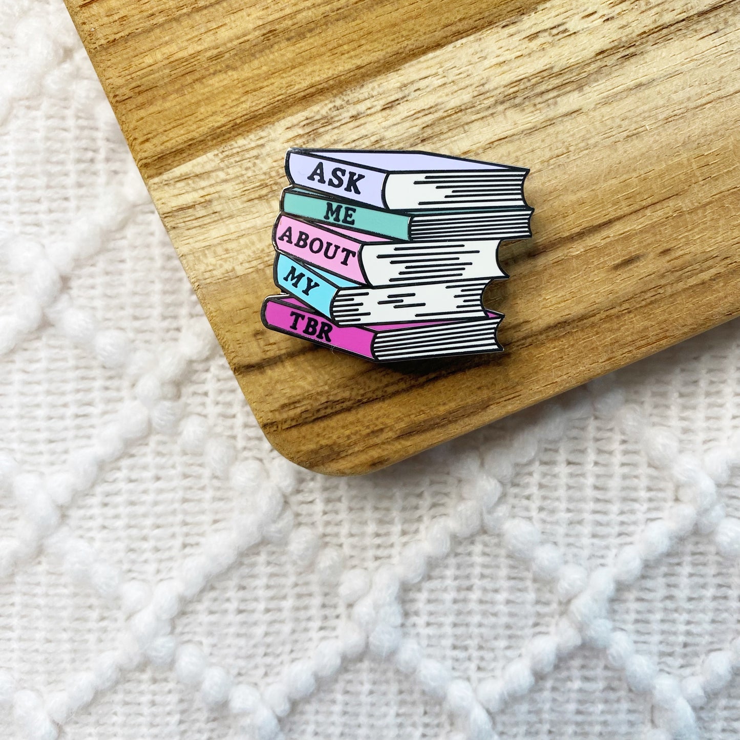 An enamel pin shaped like a stack of colorful books with the words 'ASK ME ABOUT MY TBR' in black lettering, placed on a wooden board over a white textured knit surface.