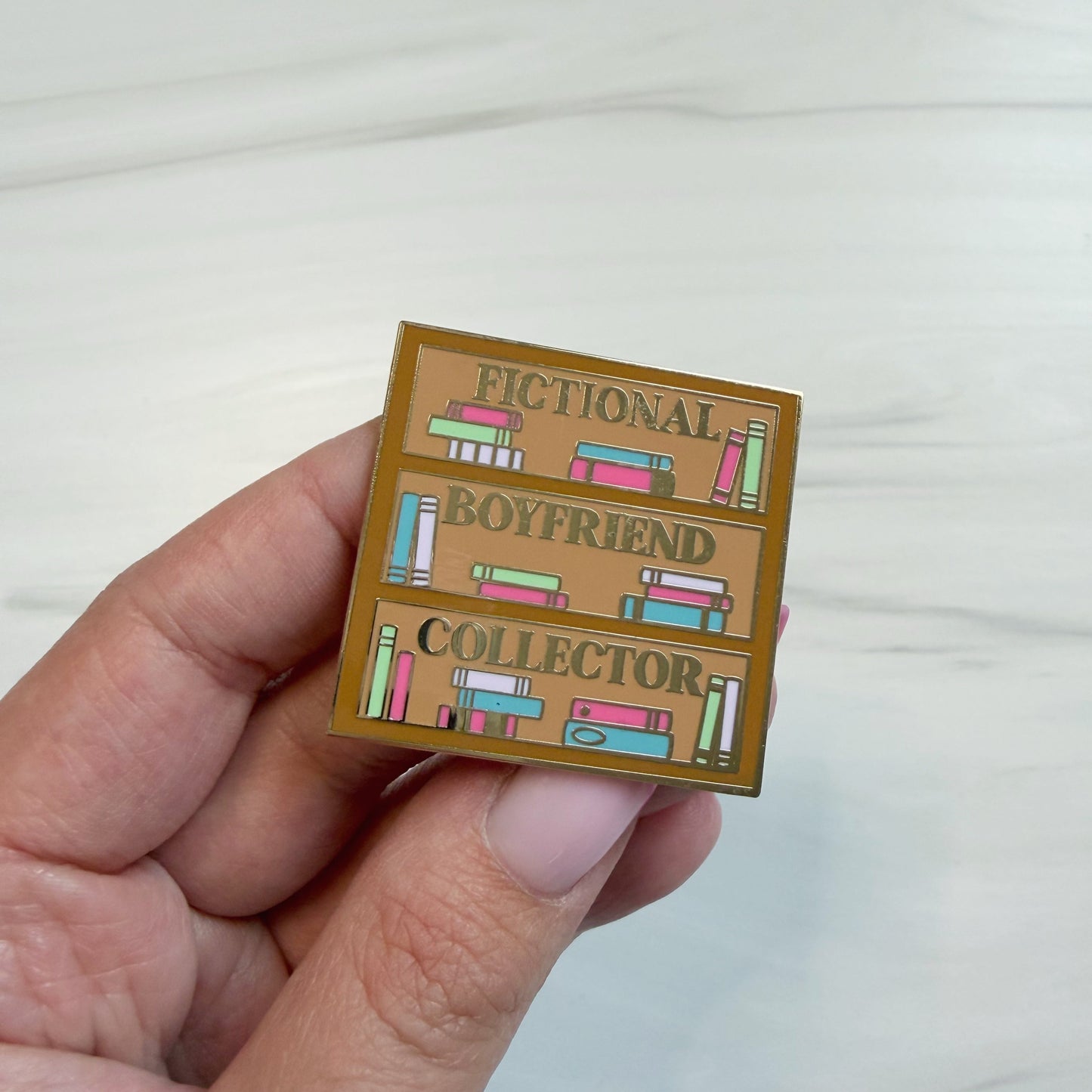 A hand holding an enamel pin featuring a bookshelf with three shelves with colorful books and the words 'FICTIONAL BOYFRIEND COLLECTOR' on a light brown background.