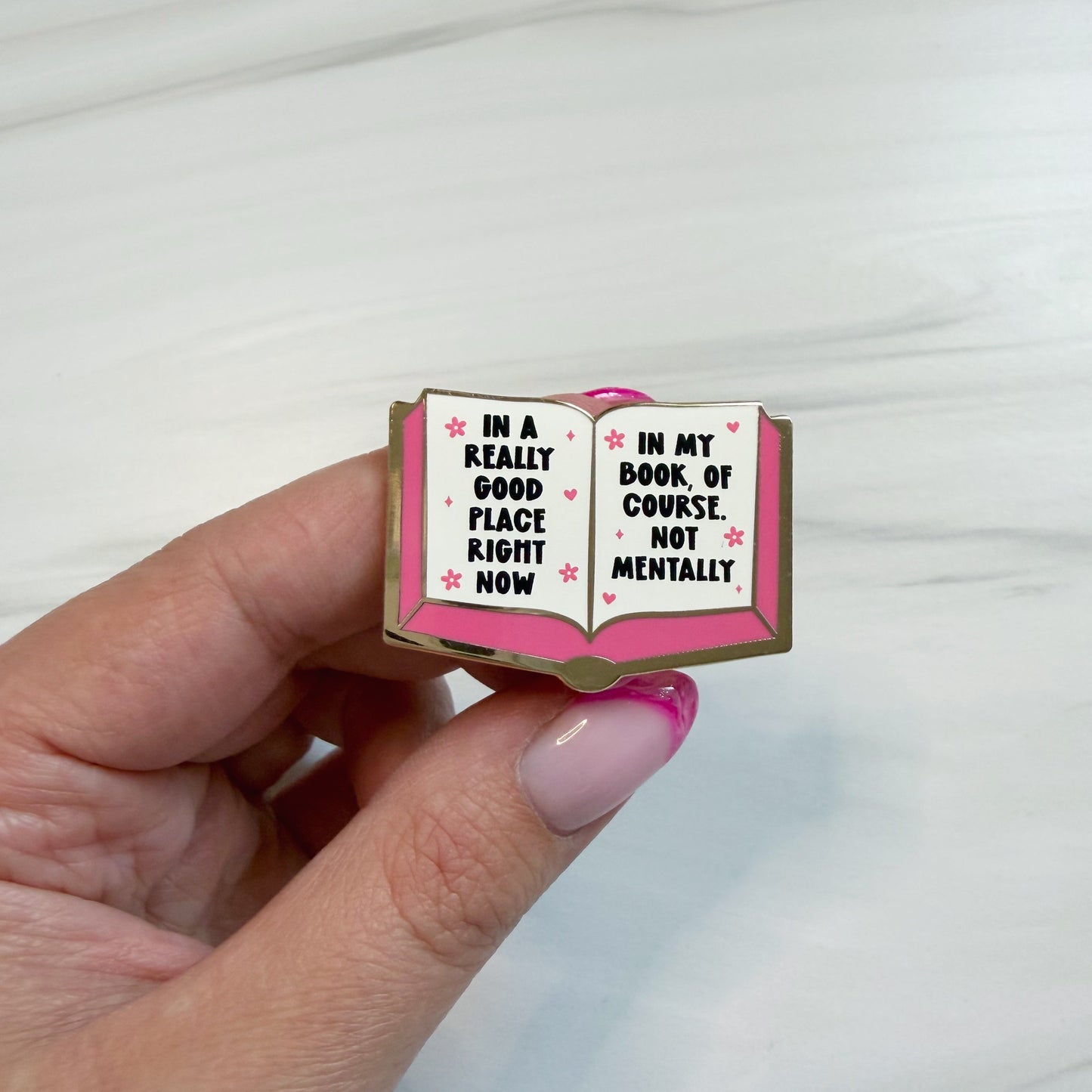 A hand with pink manicured nails holds an enamel pin shaped like an open book. The pin has a gold outline, a pink cover, and white pages with black text that reads, 'IN A REALLY GOOD PLACE RIGHT NOW' on the left page and 'IN MY BOOK OF COURSE, NOT MENTALLY' on the right page, accented with small pink hearts.