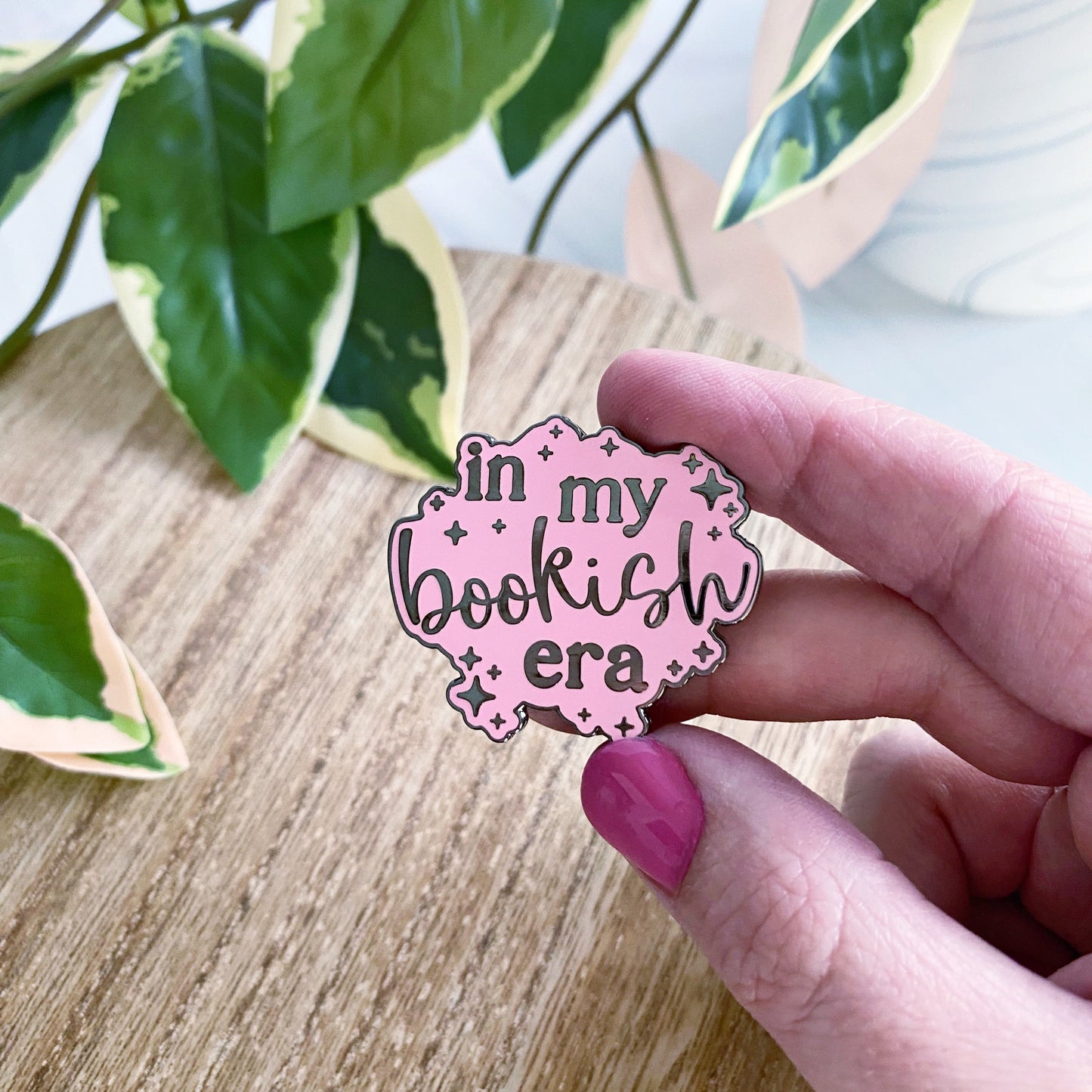 A hand with pink nail polish holds a pink enamel pin that reads 'in my bookish era' against a background of a wooden table and green variegated houseplant leaves.