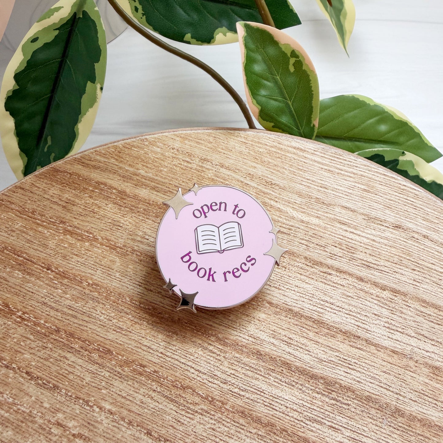 A round pink enamel pin with the screen printed text 'open to book recs' and an open book icon, resting on a wooden surface with green variegated plant leaves in the background.