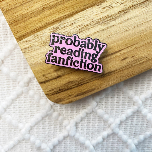 An enamel pin with the phrase 'probably reading fanfiction' in black lettering on a pink background with a black outline, resting on a wooden board over a white textured knit surface.