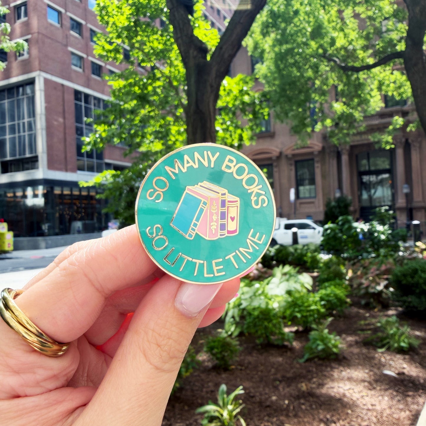 A hand holding a round enamel pin that reads 'SO MANY BOOKS SO LITTLE TIME' with a teal background and illustration of books, set against an outdoor city garden background.
