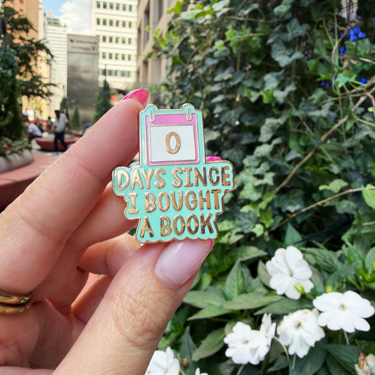 A hand holds a colorful enamel pin that reads '0 DAYS SINCE I BOUGHT A BOOK' against a blurred outdoor background of green foliage and city buildings.