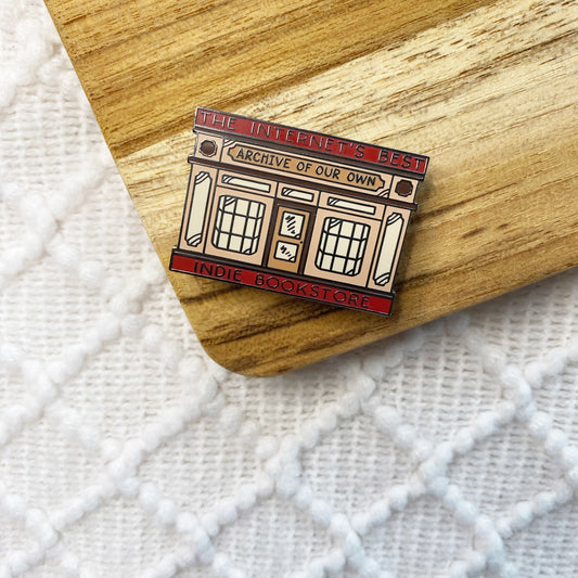 An enamel pin shaped like a bookstore storefront, placed on a wooden slice over a white textured fabric background. The pin reads “THE INTERNET’S BEST INDIE BOOKSTORE" and ARCHIVE OF OUR OWN”.