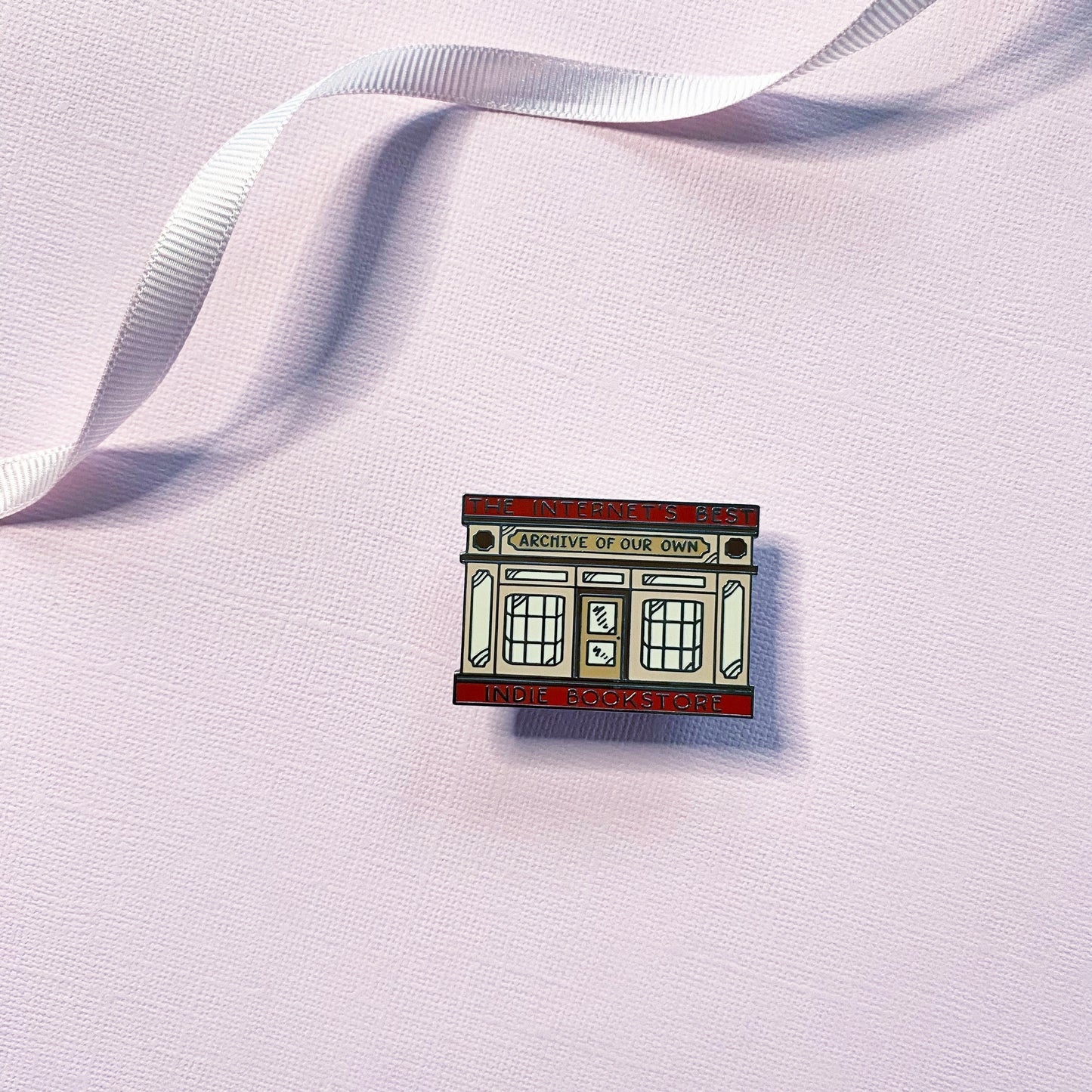 An enamel pin shaped like a bookstore storefront, with text that reads “THE INTERNET’S BEST INDIE BOOKSTORE" and ARCHIVE OF OUR OWN”, on a pale pink textured background with a white ribbon.