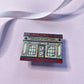 A close view of an enamel pin shaped like a bookstore storefront, with text that reads “THE INTERNET’S BEST INDIE BOOKSTORE" and ARCHIVE OF OUR OWN”, placed on a pale pink textured fabric with a white ribbon in the background.