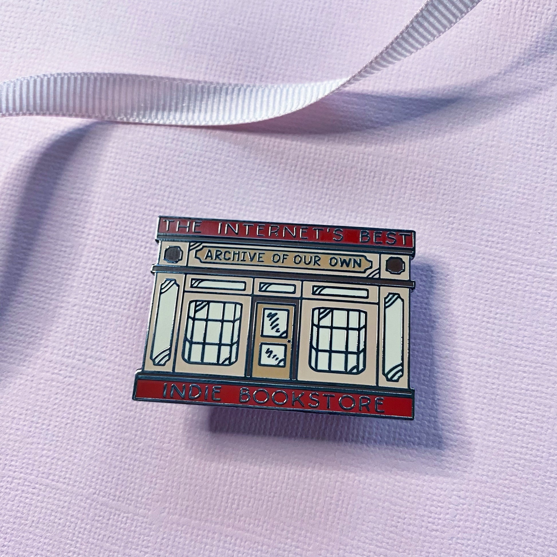 A close view of an enamel pin shaped like a bookstore storefront, with text that reads “THE INTERNET’S BEST INDIE BOOKSTORE" and ARCHIVE OF OUR OWN”, placed on a pale pink textured fabric with a white ribbon in the background.