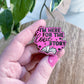 A hand with pink nail polish holds an enamel pin that reads 'I'M HERE FOR THE LOVE STORY' with an open book and heart details, set against a background of a wooden table and a green houseplant.