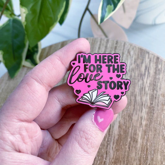 A hand with pink nail polish holds an enamel pin that reads 'I'M HERE FOR THE LOVE STORY' with an open book and heart details, set against a background of a wooden table and a green houseplant.