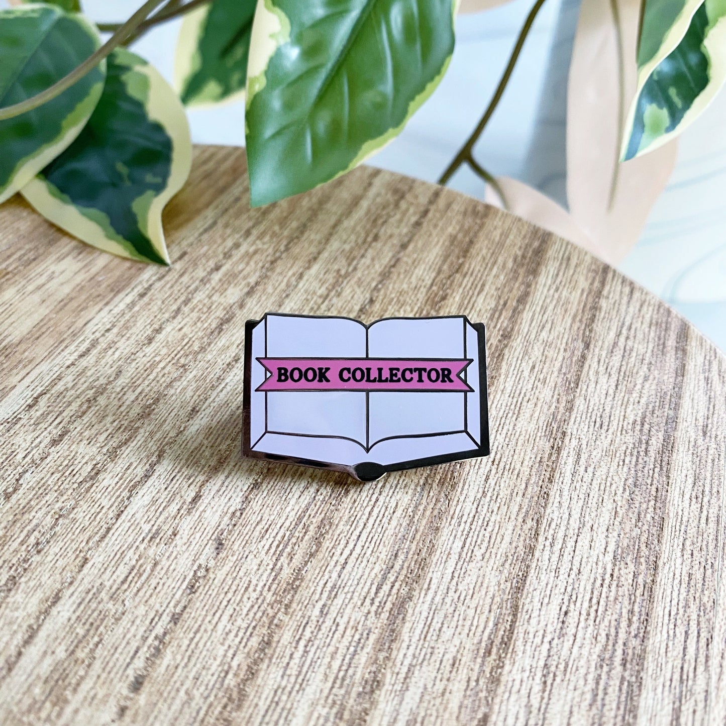 An enamel pin shaped like an open book with purple pages and a pink banner reading “BOOK COLLECTOR,” placed on a wooden surface with green variegated leaves in the background.