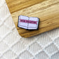 An enamel pin shaped like an open book with a pink banner reading “BOOK COLLECTOR,” placed on a wooden surface over a white textured knit fabric.