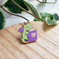 An enamel pin shaped like a green dragon reading a purple book with a gold heart, resting on a light wood surface with green leaves in the background.