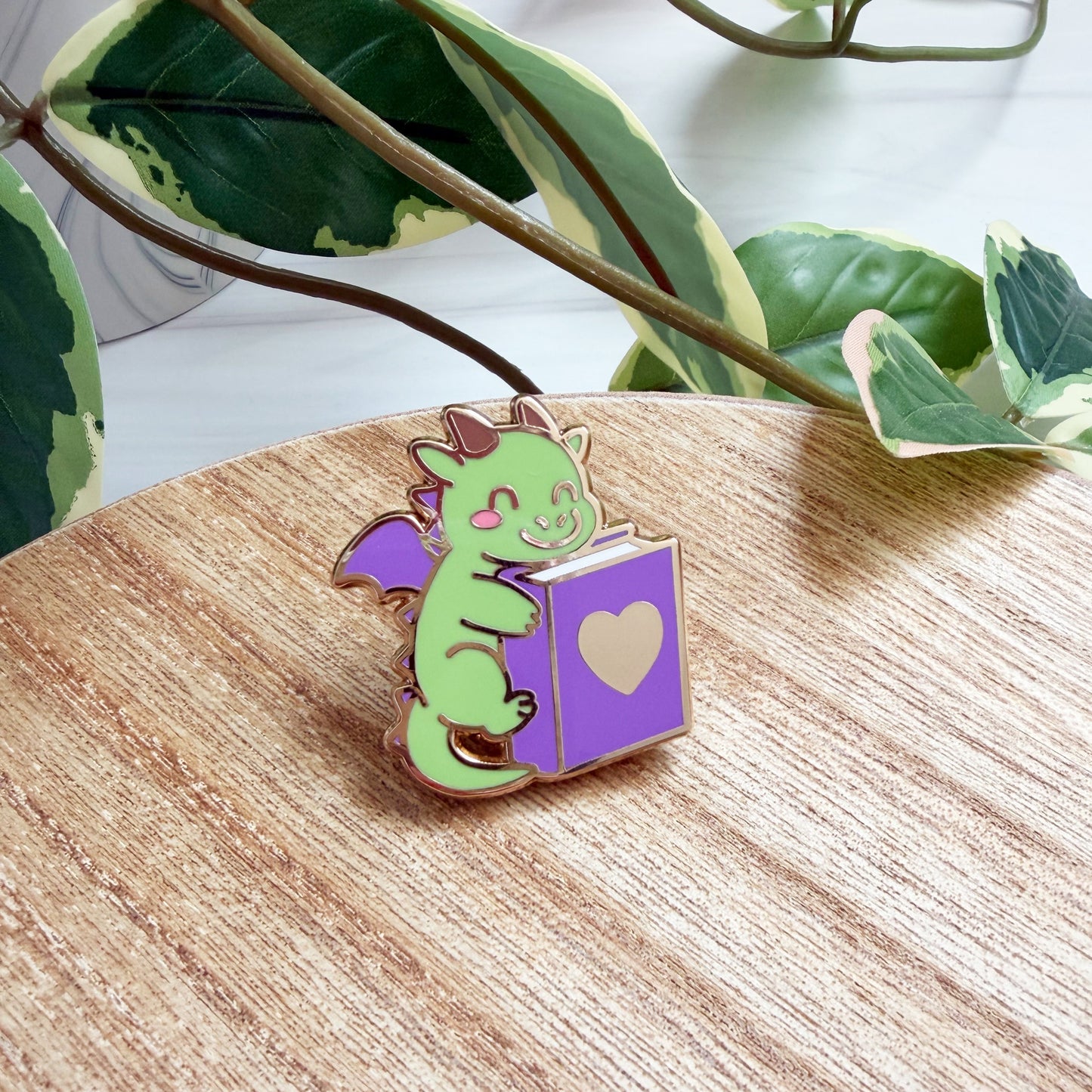 An enamel pin shaped like a green dragon reading a purple book with a gold heart, resting on a light wood surface with green leaves in the background.