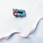 A small enamel pin shaped like a stack of colorful books next to a pink teacup and saucer, with the words "books & tea" above them, placed on a white textured surface with a pale pink ribbon running diagonally across the frame.