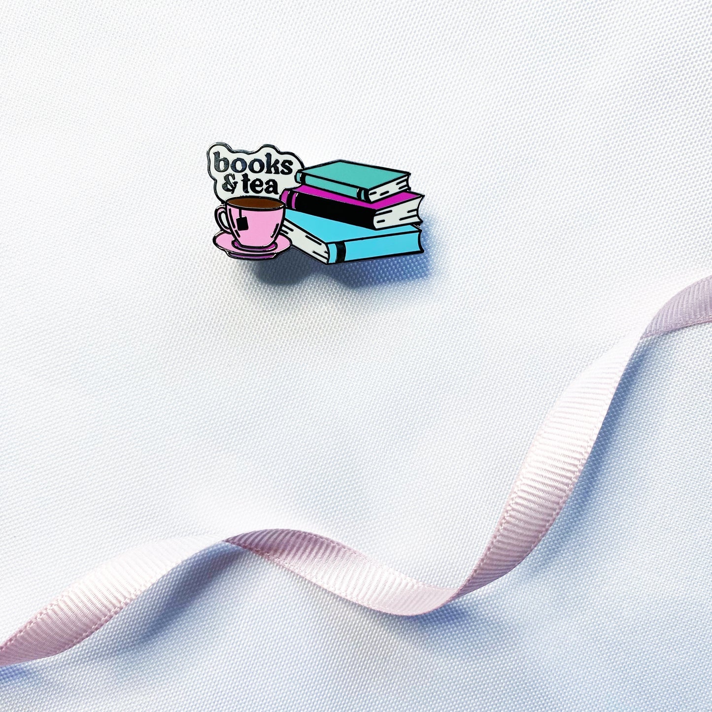 A small enamel pin shaped like a stack of colorful books next to a pink teacup and saucer, with the words "books & tea" above them, placed on a white textured surface with a pale pink ribbon running diagonally across the frame.