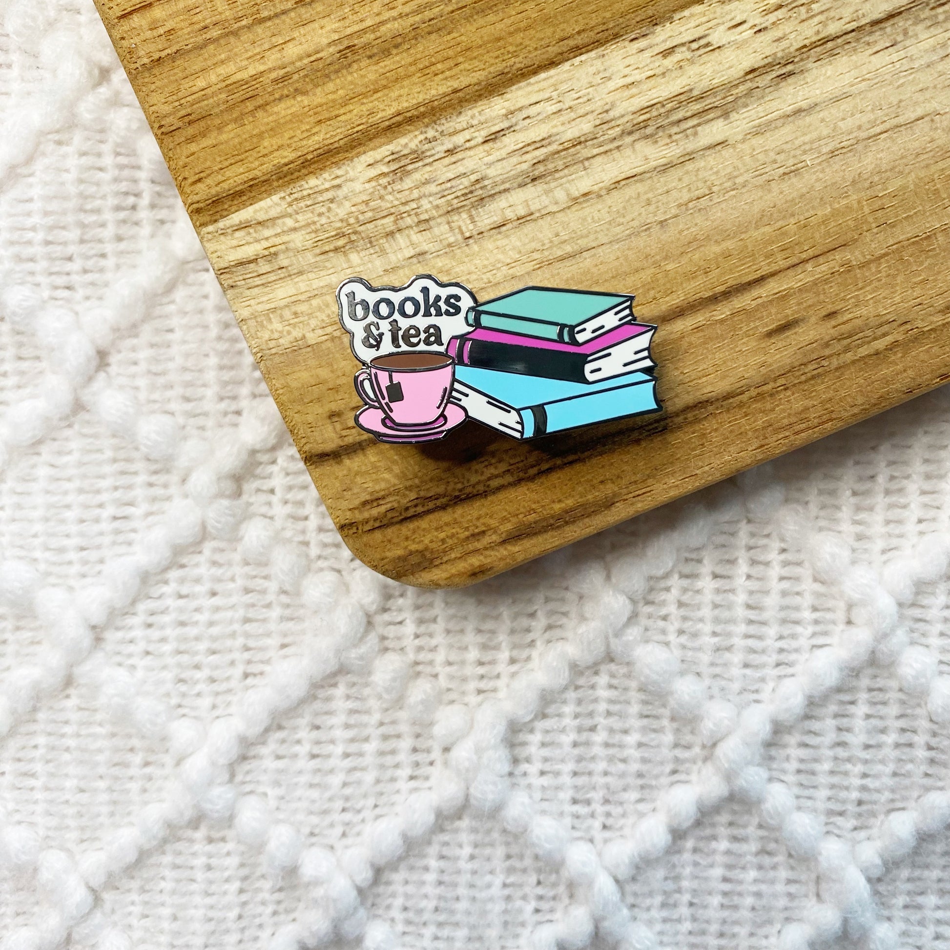 A small enamel pin shaped like a stack of books and a pink teacup saucer with the words “books & tea,” placed on a wooden board over a white textured knit surface.