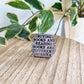 An enamel pin with the text 'BUYING BOOKS AND READING BOOKS ARE DIFFERENT HOBBIES' in black lettering on a purple background, resting on a light wood tabletop with green variegated houseplants in the background.