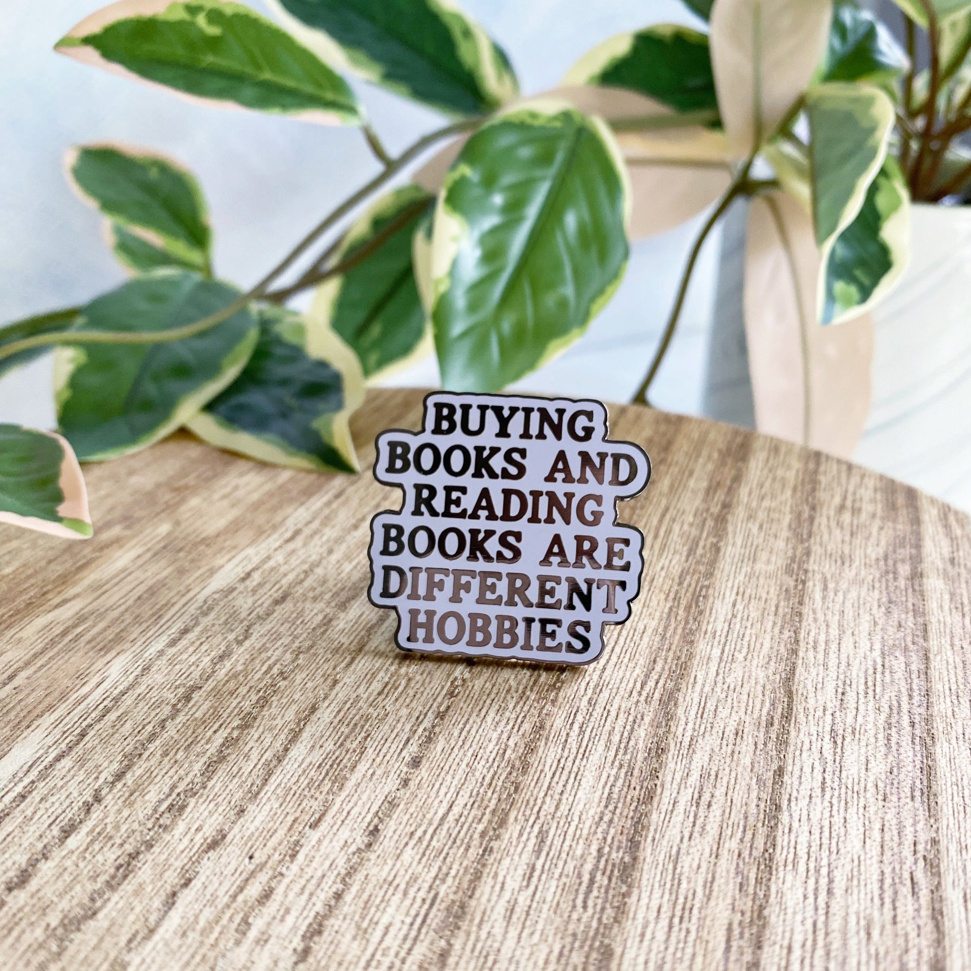 An enamel pin with the text 'BUYING BOOKS AND READING BOOKS ARE DIFFERENT HOBBIES' in black lettering on a purple background, resting on a light wood tabletop with green variegated houseplants in the background.