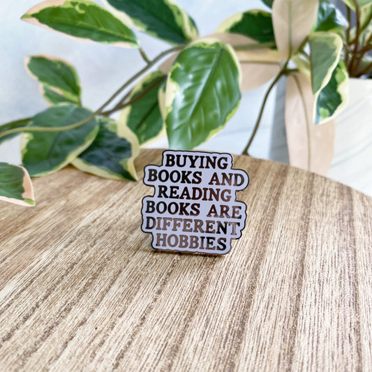 An enamel pin with the text 'BUYING BOOKS AND READING BOOKS ARE DIFFERENT HOBBIES' in black lettering on a purple background, resting on a light wood tabletop with green variegated houseplants in the background.