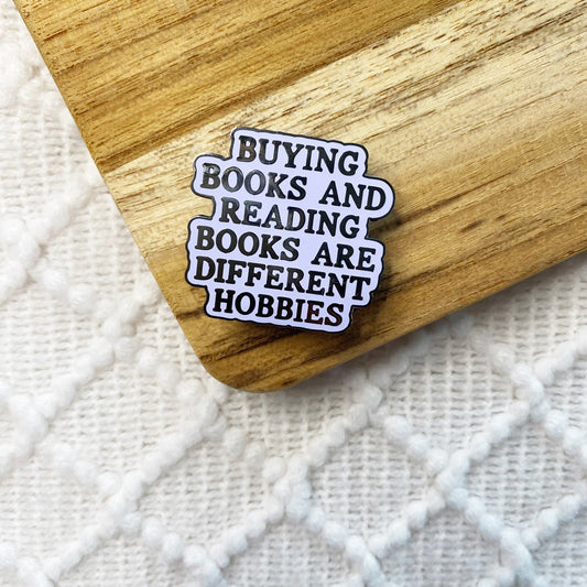 An enamel pin with the text 'BUYING BOOKS AND READING BOOKS ARE DIFFERENT HOBBIES' in black lettering on a light purple background with a black outline, resting on a wooden surface over a white textured knit fabric.