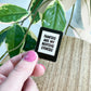 A hand with pink nail polish holds an enamel pin shaped like an e-reader or tablet with the text 'FANFICS ARE MY BEDTIME STORIES' in black letters on a white background. The pin is shown against a background of green houseplants and a wooden tabletop.