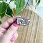 A hand with pink nail polish holds an enamel pin shaped like a stack of books and a coffee cup, with the text ' fueled by books and coffee', against a wooden table with green variegated leaves in the background.