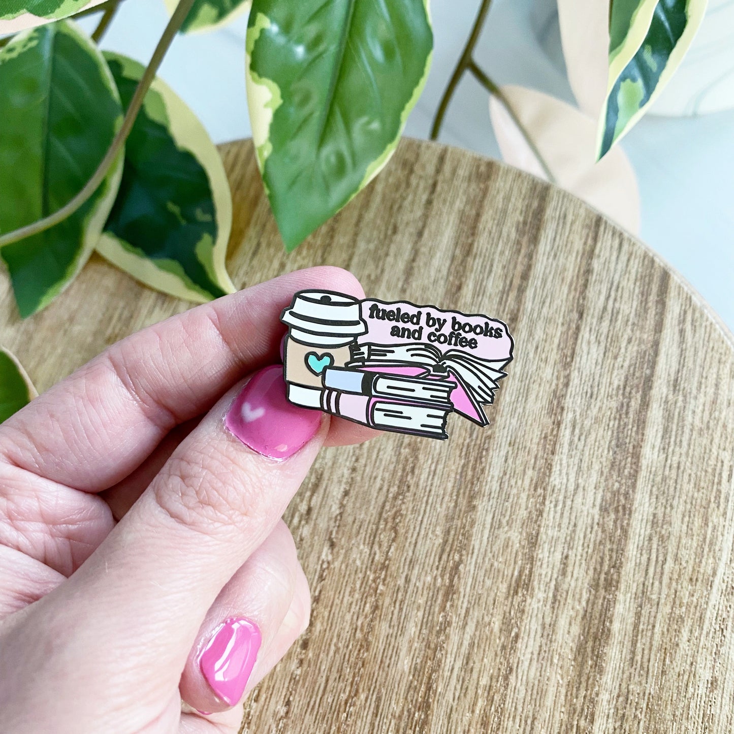A hand with pink nail polish holds an enamel pin shaped like a stack of books and a coffee cup, with the text ' fueled by books and coffee', against a wooden table with green variegated leaves in the background.