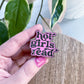 A hand with pink nail polish holds an enamel pin that reads 'hot girls read' in black lettering with a light pink outline, positioned over a wooden surface with green houseplant leaves in the background.