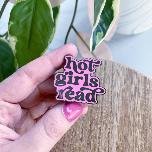 A hand with pink nail polish holds an enamel pin that reads 'hot girls read' in black lettering with a light pink outline, positioned over a wooden surface with green houseplant leaves in the background.