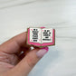 A hand with pink manicured nails holds an enamel pin shaped like an open book. The pin has a gold outline, a pink cover, and white pages with black text that reads, 'IN A REALLY GOOD PLACE RIGHT NOW' on the left page and 'IN MY BOOK OF COURSE, NOT MENTALLY' on the right page, accented with small pink hearts.