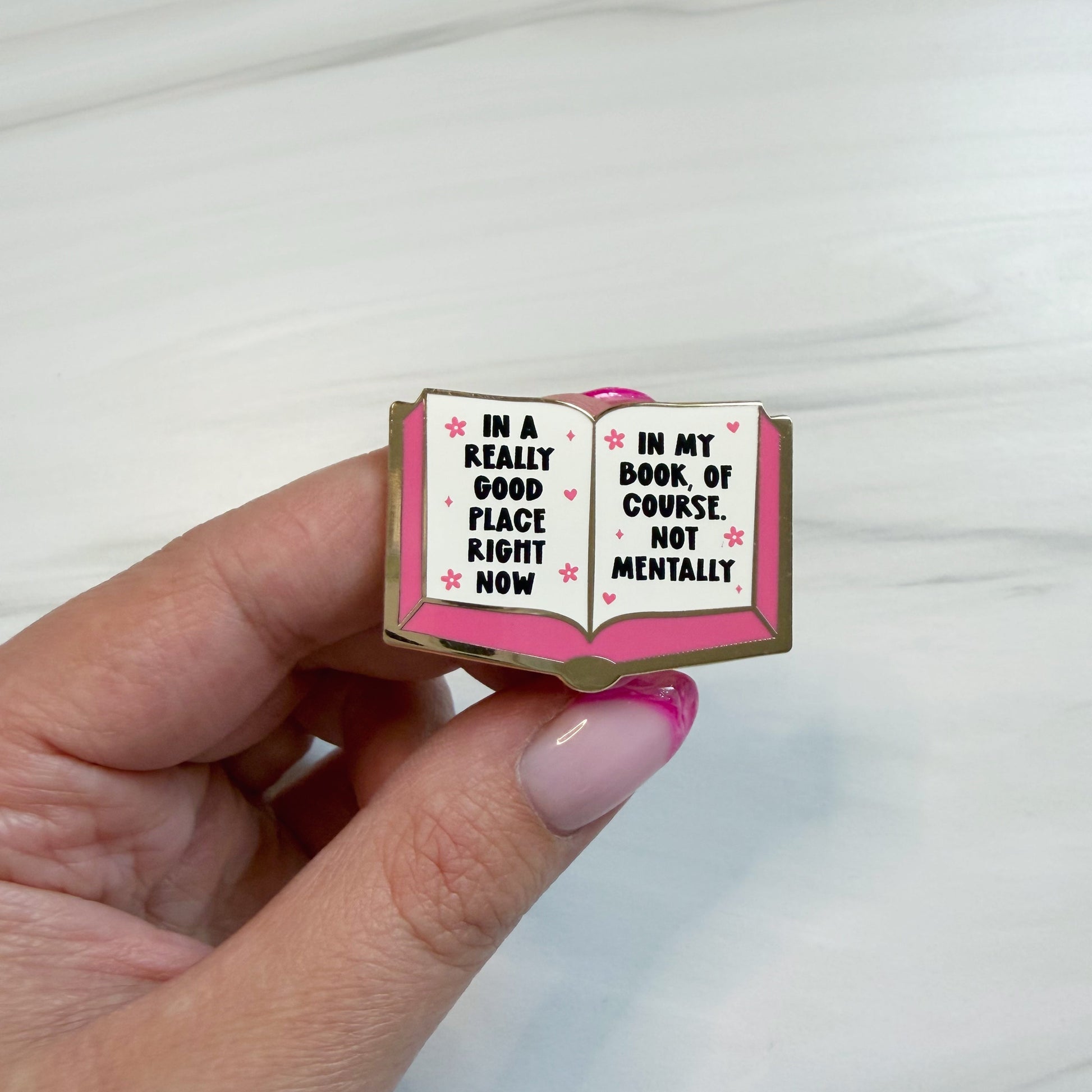 A hand with pink manicured nails holds an enamel pin shaped like an open book. The pin has a gold outline, a pink cover, and white pages with black text that reads, 'IN A REALLY GOOD PLACE RIGHT NOW' on the left page and 'IN MY BOOK OF COURSE, NOT MENTALLY' on the right page, accented with small pink hearts.