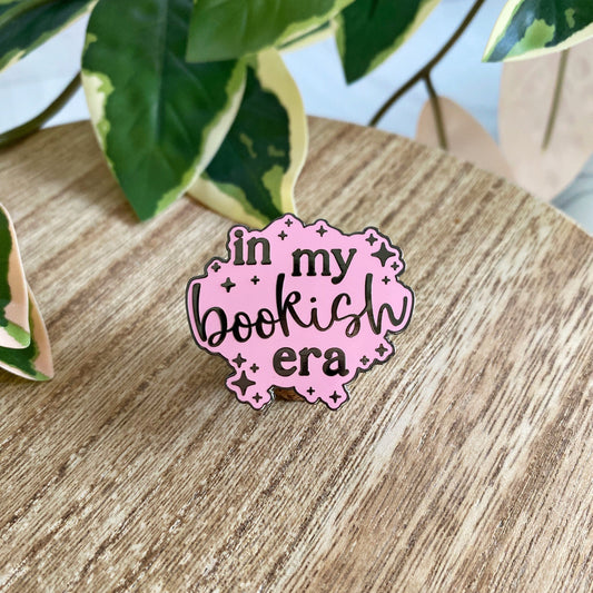 A pink enamel pin with the text 'in my bookish era' in black text, displayed on a wooden surface with green variegated plant leaves in the background.