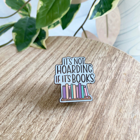 An enamel pin with the phrase “IT’S NOT HOARDING IF IT’S BOOKS” above a row of colorful books, displayed on a round wooden surface with blurred green leaves in the background.