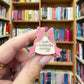 A hand holding an enamel pin shaped like a pink bow and ribbon, with the words 'main character energy' written on it, set against a blurred background of bookshelves.