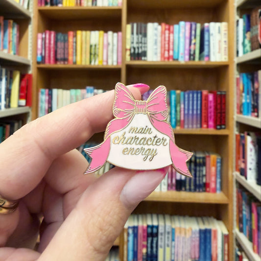 A hand holding an enamel pin shaped like a pink bow and ribbon, with the words 'main character energy' written on it, set against a blurred background of bookshelves.