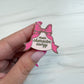 A hand holding a pink enamel pin shaped like a bow and banner that reads 'main character energy' in gold script, against a light marble background.