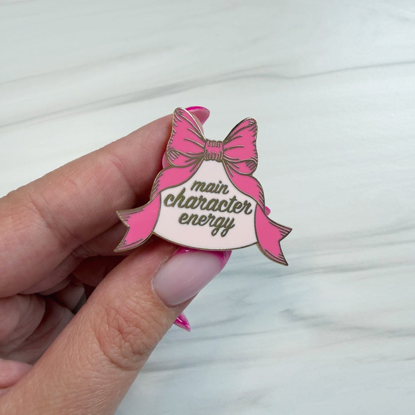 A hand holding a pink enamel pin shaped like a bow and banner that reads 'main character energy' in gold script, against a light marble background.