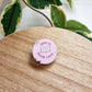 A round pink enamel pin with the screen printed text 'open to book recs' and an open book icon, resting on a wooden surface with green variegated plant leaves in the background.
