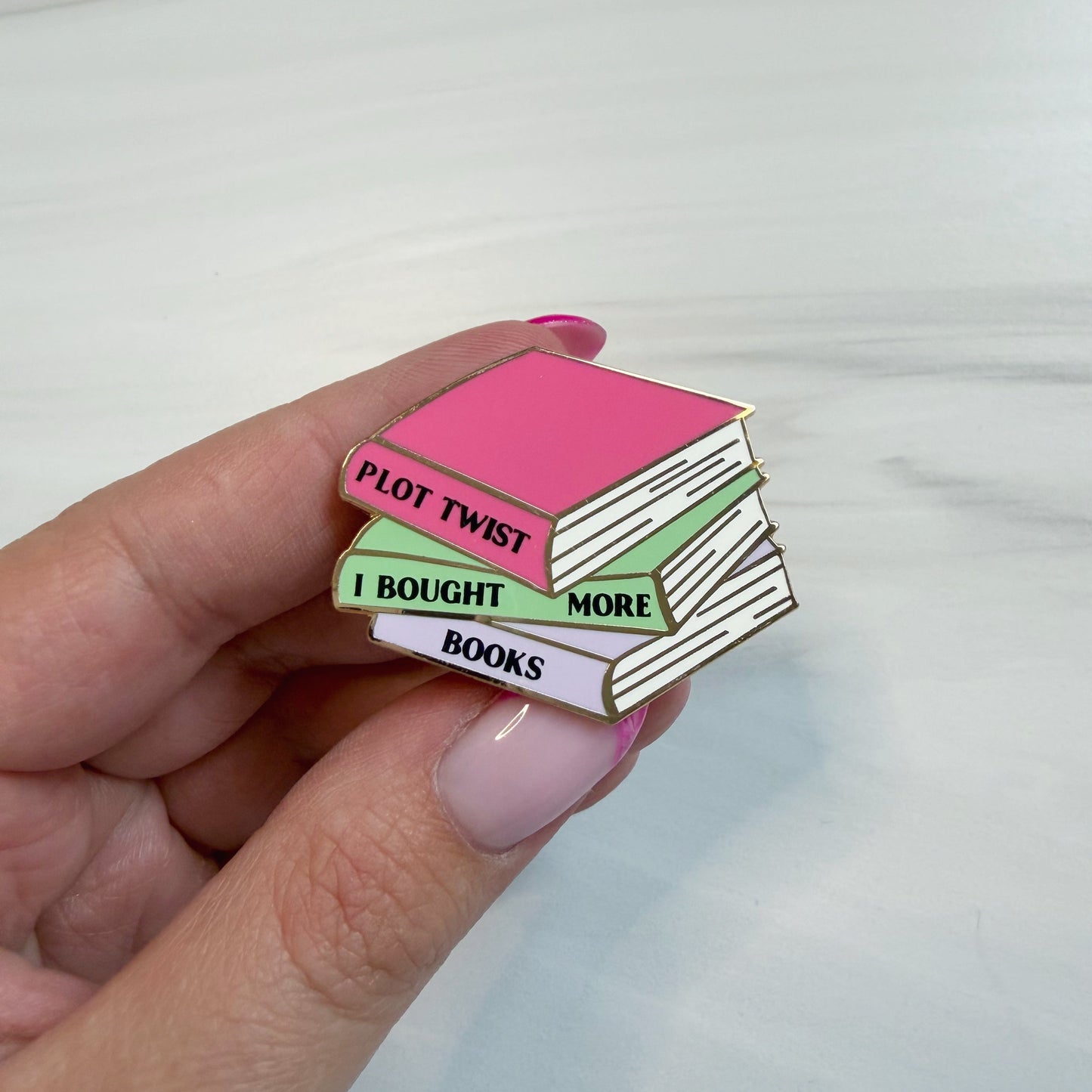 A hand with pink manicured nails holds an enamel pin shaped like a stack of three books. The top book is pink with the words 'PLOT TWIST' in black, the middle is light green with 'I BOUGHT MORE' in black, and the bottom is lavender with 'BOOKS' in black.