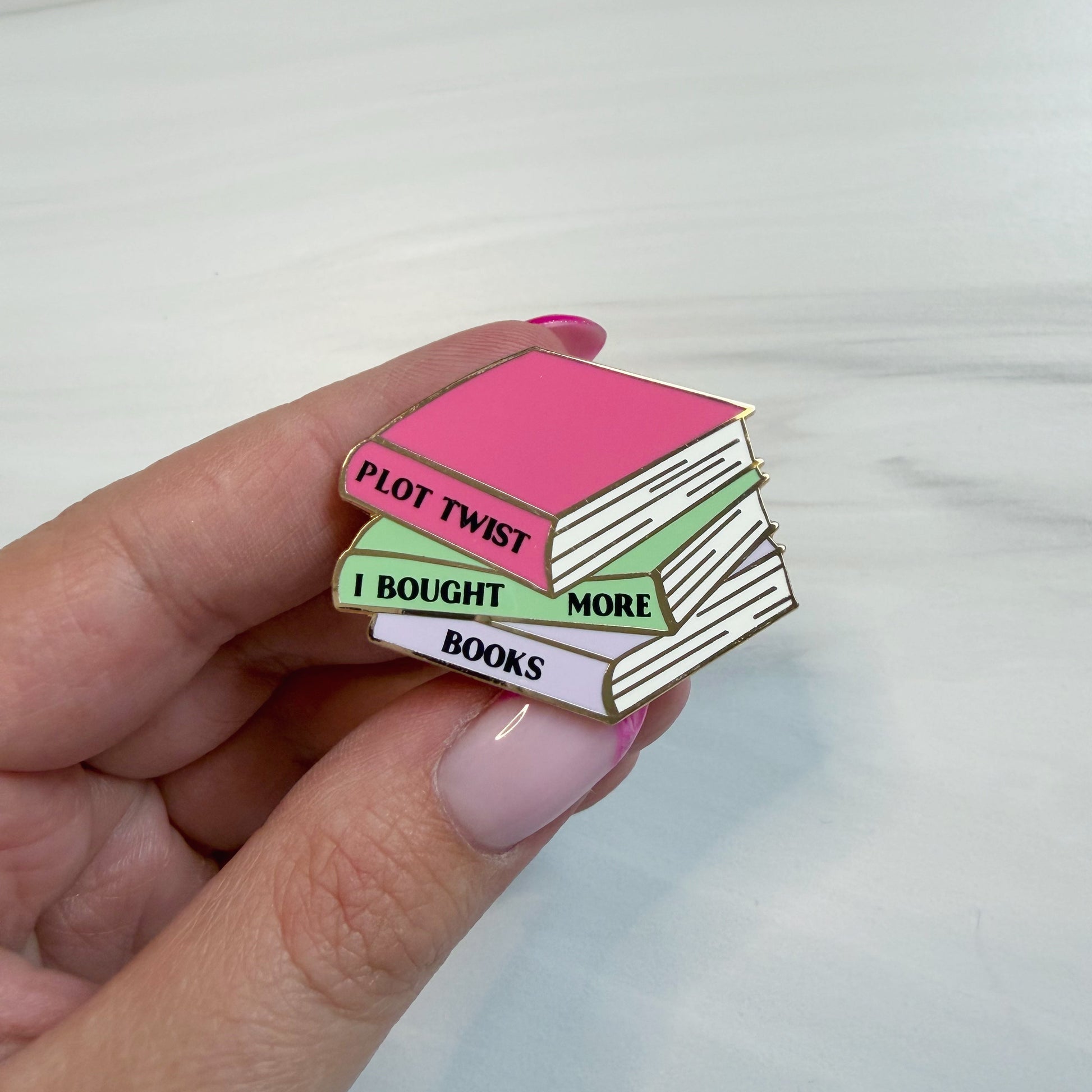 A hand with pink manicured nails holds an enamel pin shaped like a stack of three books. The top book is pink with the words 'PLOT TWIST' in black, the middle is light green with 'I BOUGHT MORE' in black, and the bottom is lavender with 'BOOKS' in black.