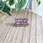 An enamel pin with the phrase 'probably reading fanfiction' in black lettering on a pink background, placed on a light wood tabletop with green variegated leaves in the background.