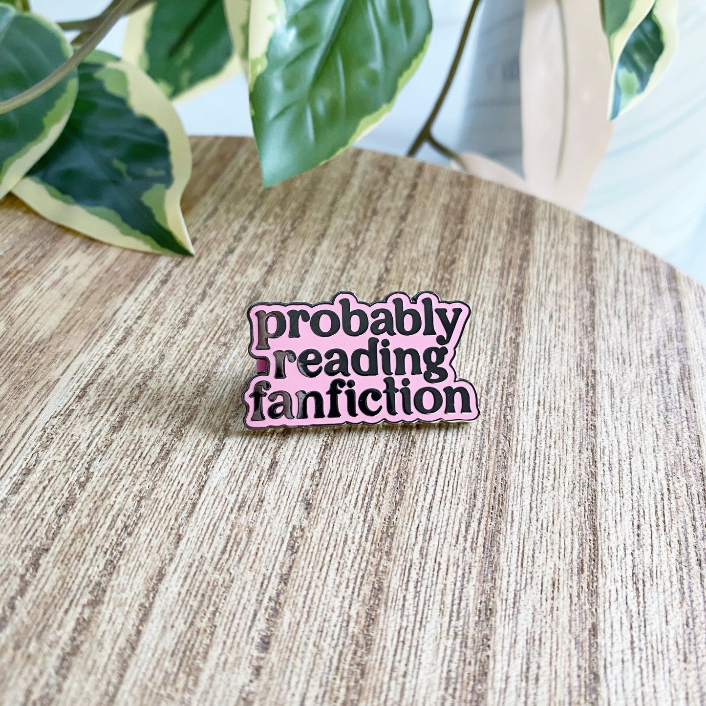 An enamel pin with the phrase 'probably reading fanfiction' in black lettering on a pink background, placed on a light wood tabletop with green variegated leaves in the background.