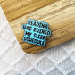 An enamel pin with the text 'READING HAS RUINED MY SLEEP SCHEDULE' in black letters on a light blue background, resting on a wooden surface over a white textured blanket.