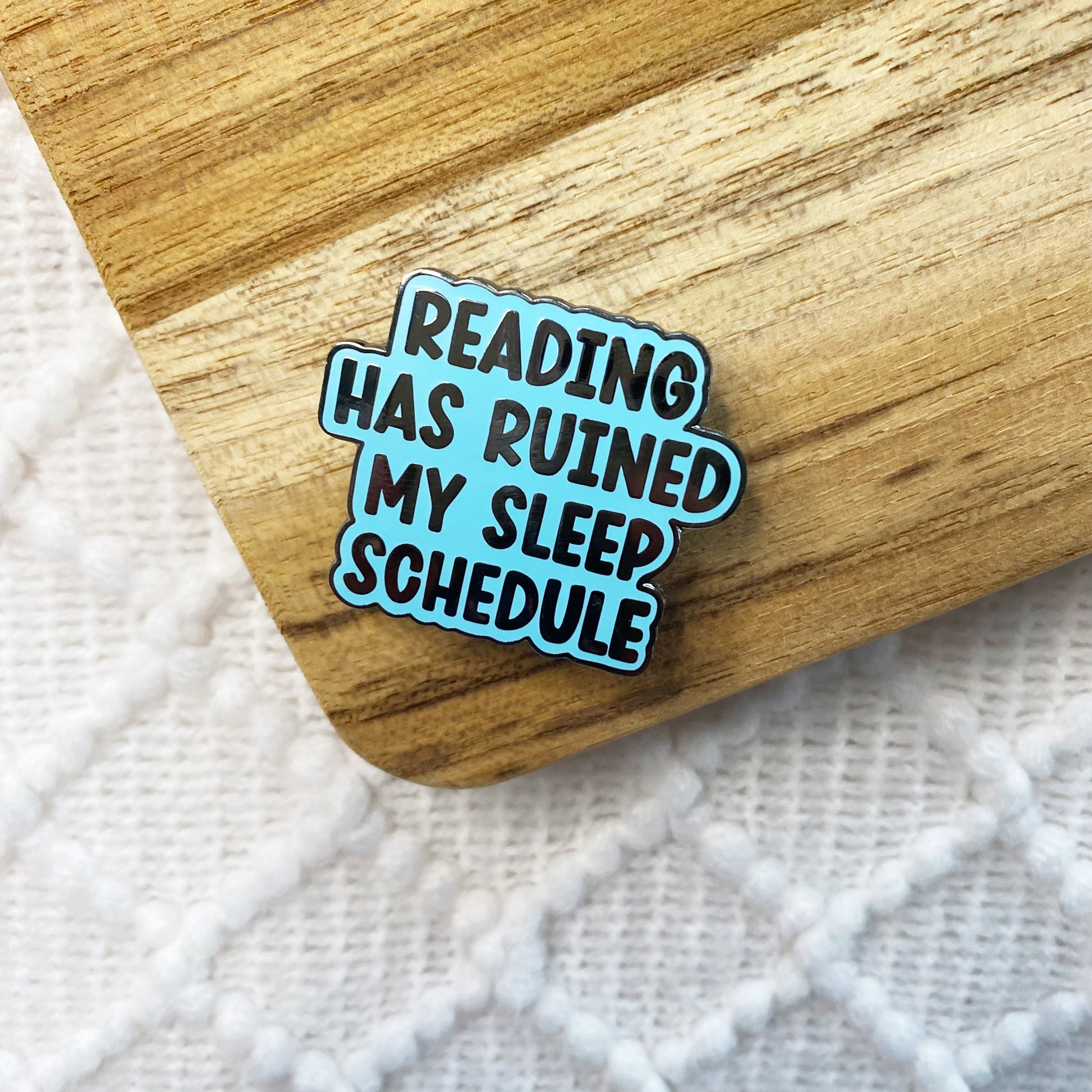 An enamel pin with the text 'READING HAS RUINED MY SLEEP SCHEDULE' in black letters on a light blue background, resting on a wooden surface over a white textured blanket.