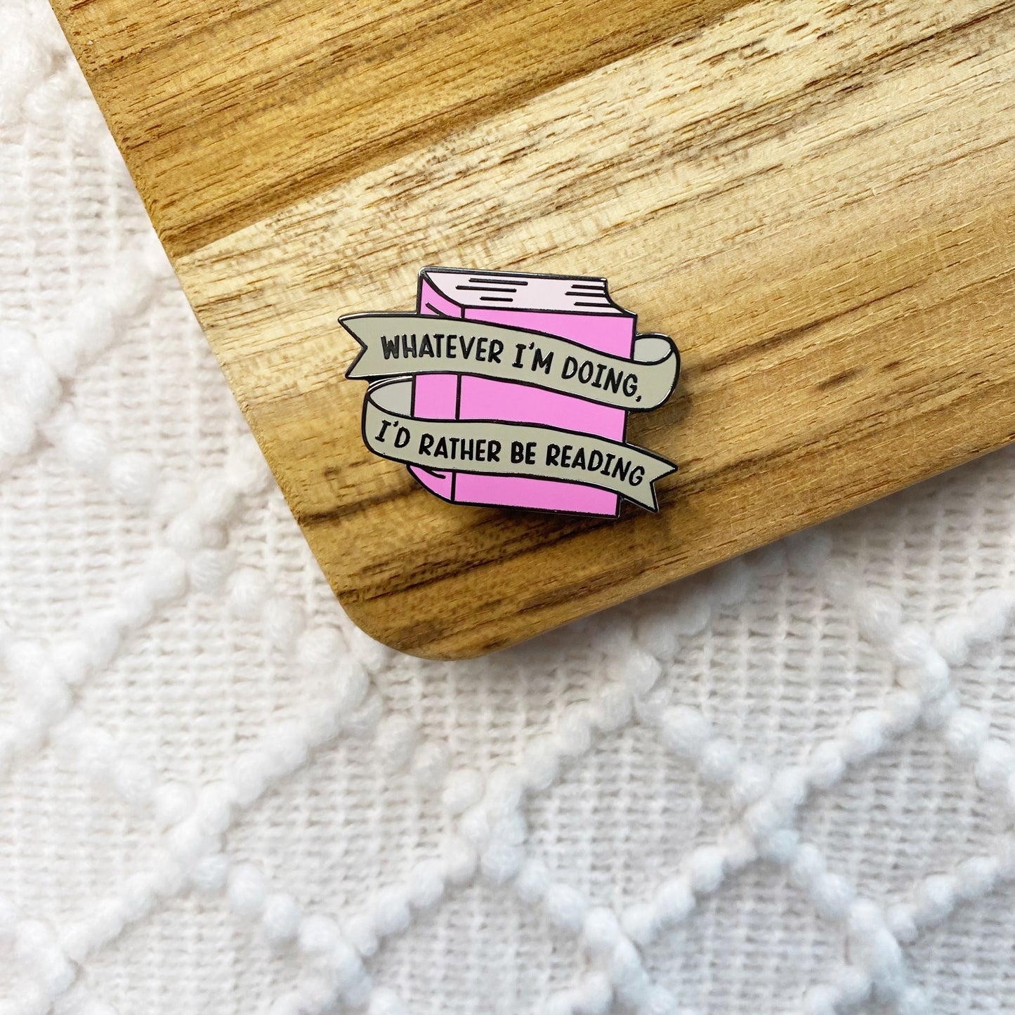 A pink enamel pin shaped like a closed book with the text 'WHATEVER I'M DOING, I'D RATHER BE READING' in two banners, placed on a wooden surface over a white textured fabric background.