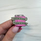 A hand with pink manicured nails holds an enamel pin shaped like a pink closed book with two banners reading, 'WHATEVER I'M DOING, I'D RATHER BE READING.' The pin is shown against a light marble-like background.